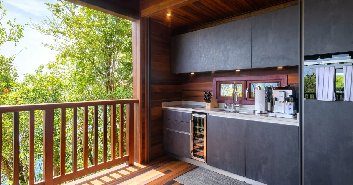 PHOTO — outdoor kitchen with dark cabinetry and wine fridge, opening onto the deck and rainforest. 1960x1026