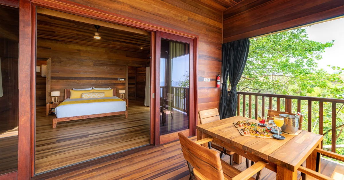 PHOTO — bedroom opening fully onto the deck, bed on the left, dining table with ice bucket on the right. 1960x1026