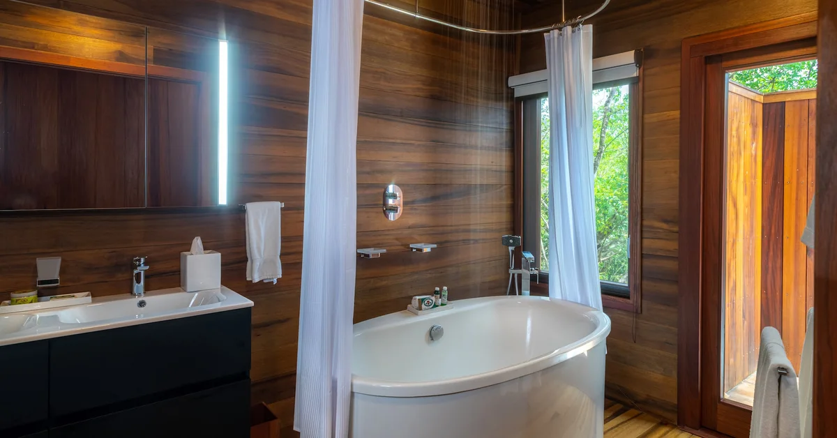 PHOTO — bathroom with freestanding soaking tub, white curtain, vanity and basin, dark timber walls, vertical