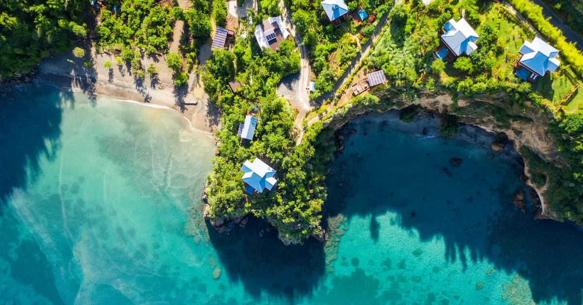 PHOTO — aerial along the coastline: rocky headland, pale beach, villas in the canopy, vivid turquoise shallows.