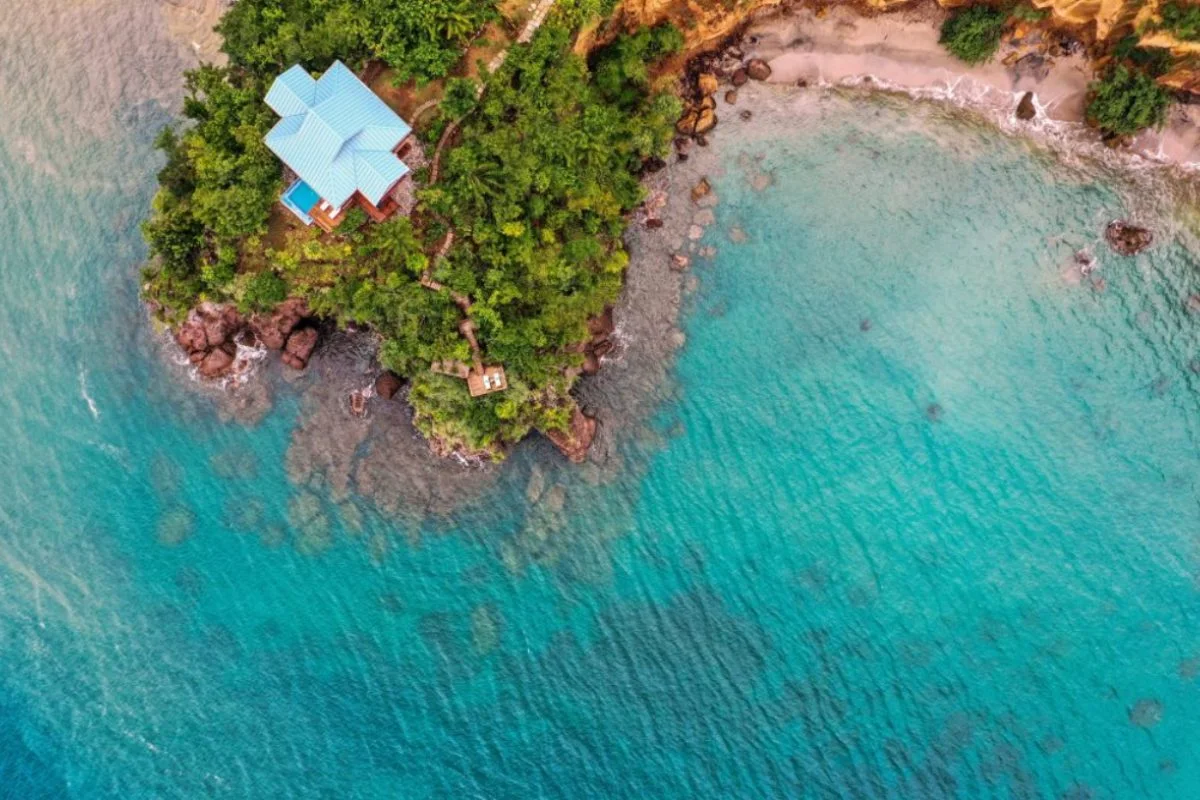 PHOTO — top-down aerial of the wooded headland with a blue-roofed villa, beach and reef in clear turquoise