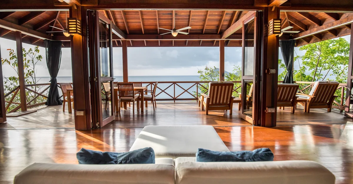 PHOTO — large open-sided living pavilion with sectional sofa, dining table and loungers under a timber roof,