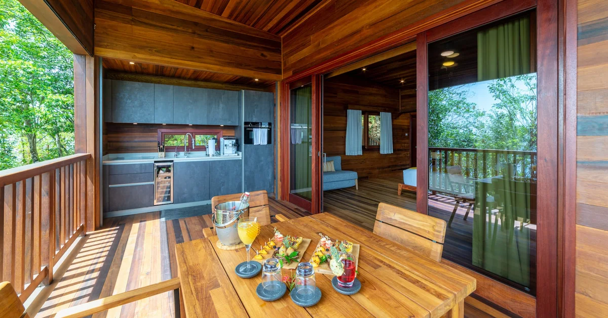PHOTO — deck dining table laid with fruit and drinks, open kitchen and bedroom behind, green railing view.